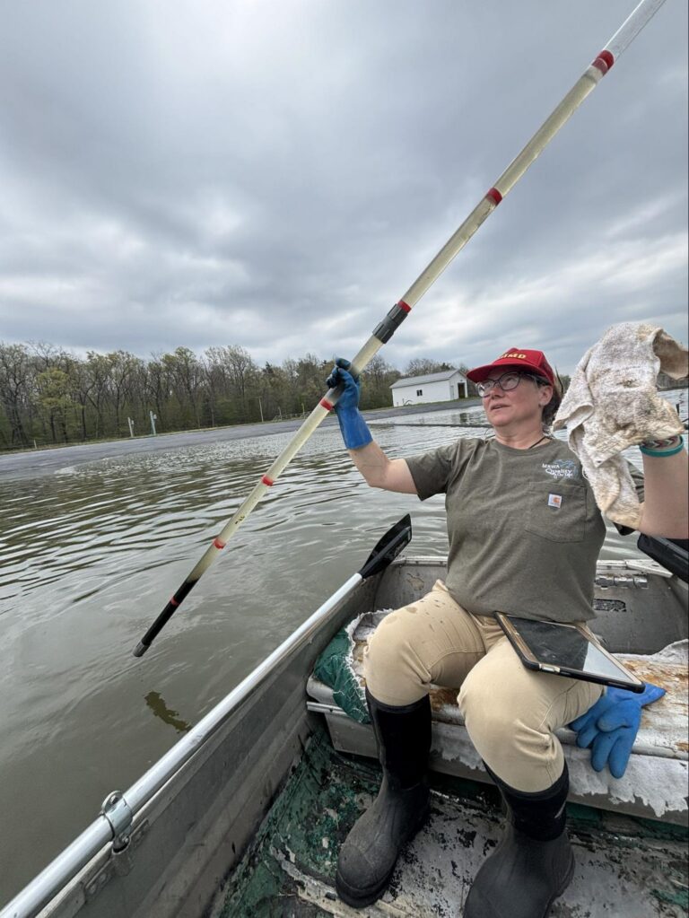 Wastewater operator in a boat measuring lagoon sludge depth with a marked survey pole.