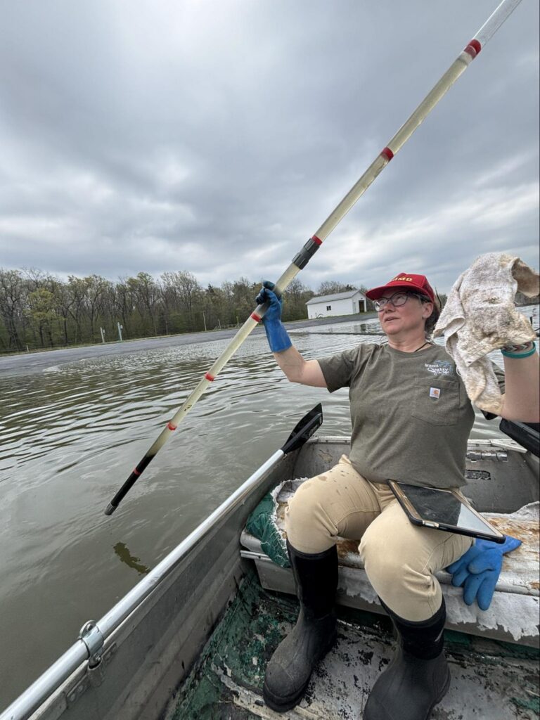 Wastewater operator in a boat measuring lagoon sludge depth with a marked survey pole.