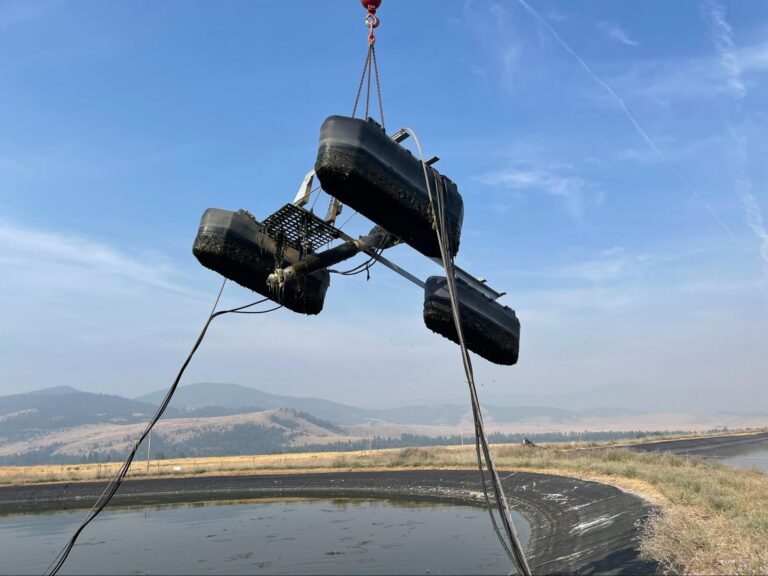 Crane lifting a sludge-coated floating aerator out of a lined wastewater lagoon for service.